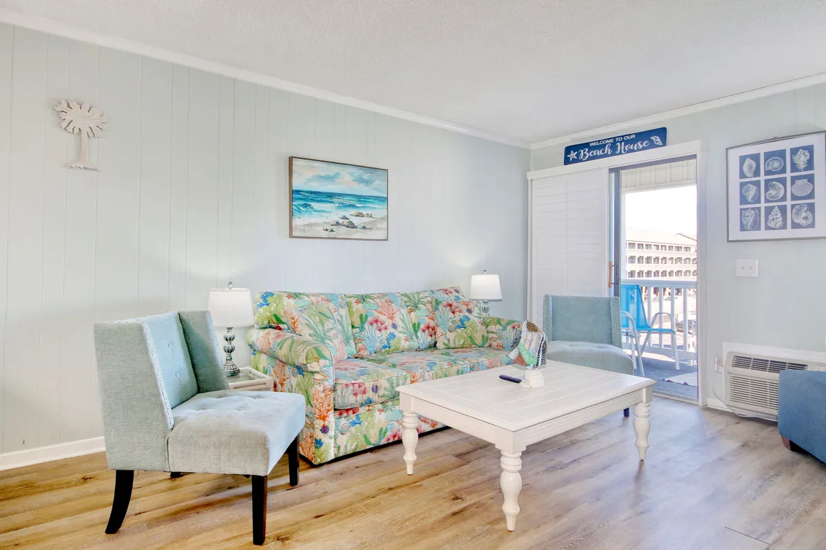 A vibrant, laid-back living area with a floral couch, coastal-themed artwork, a white coffee table, and blue accent chairs. The large sliding doors in the background suggest easy access to the balcony and the inviting coastal atmosphere beyond.