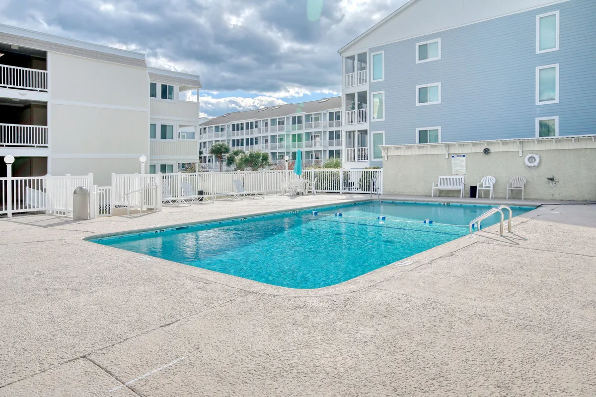 A spacious outdoor swimming pool surrounded by numerous lounge chairs, with nearby resort-style buildings visible in the background. The pool area feels inviting and is arranged for socializing, sunbathing, and relaxing with friends.