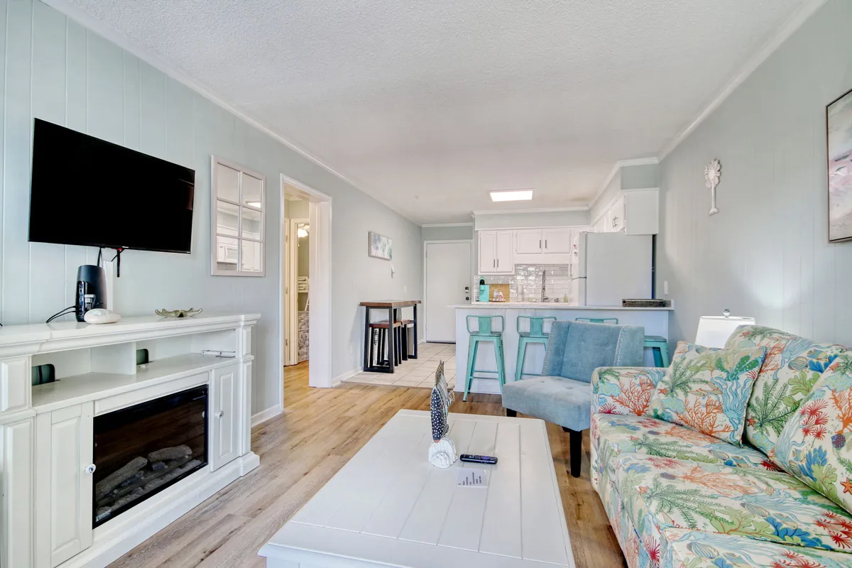 A cozy and cheerful living area that blends a modern entertainment unit, colorful floral sofa, and a kitchen island. The open layout visually connects the living room and kitchen, creating a spacious and welcoming environment.