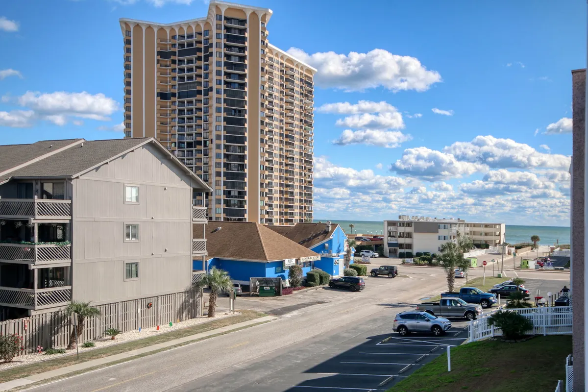 A captivating view from the building, highlighting a tall apartment structure, a blue commercial building, and a shimmering ocean in the distance. The scene radiates a relaxed beach vibe, with the ocean clearly visible as a backdrop to vibrant community life.