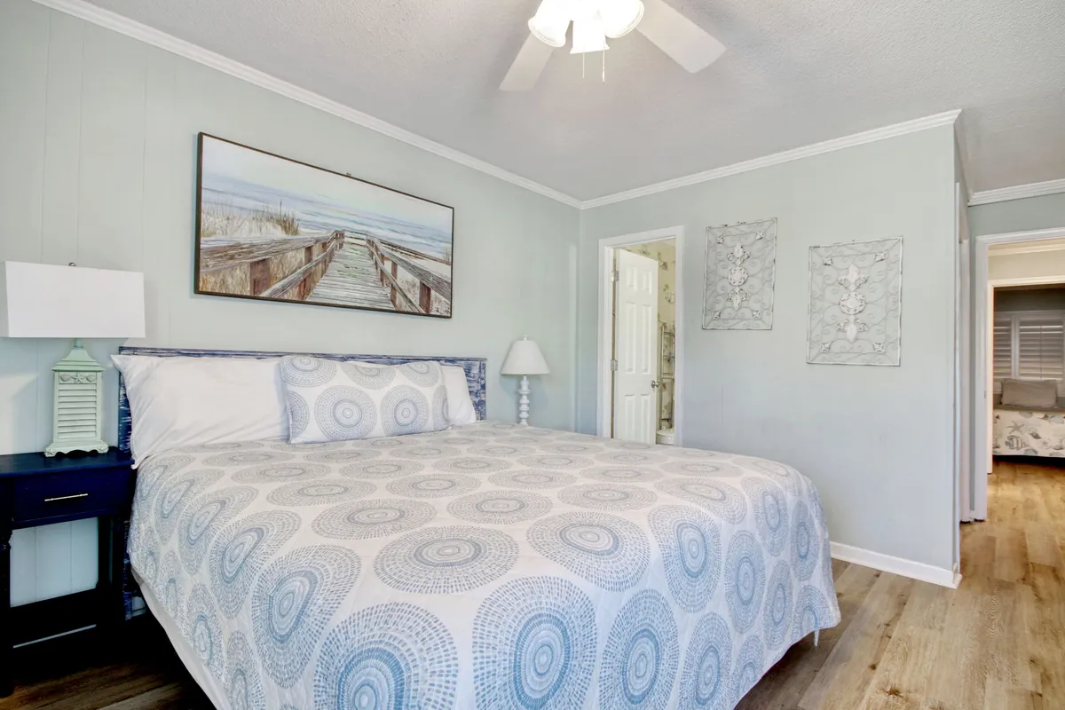 A tranquil bedroom scene with a bed dressed in patterned bedding, a framed artwork of a pier above the headboard, a bedside lamp, and a nightstand, all bathed in soft natural light.