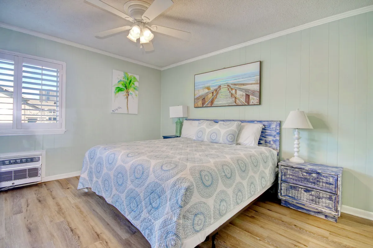 A serene bedroom featuring a bed with patterned bedding, beach-themed artwork on the wall, and a nightstand with a lamp. Sunlight gently illuminates the space, creating an inviting ambiance ideal for rest and relaxation.
