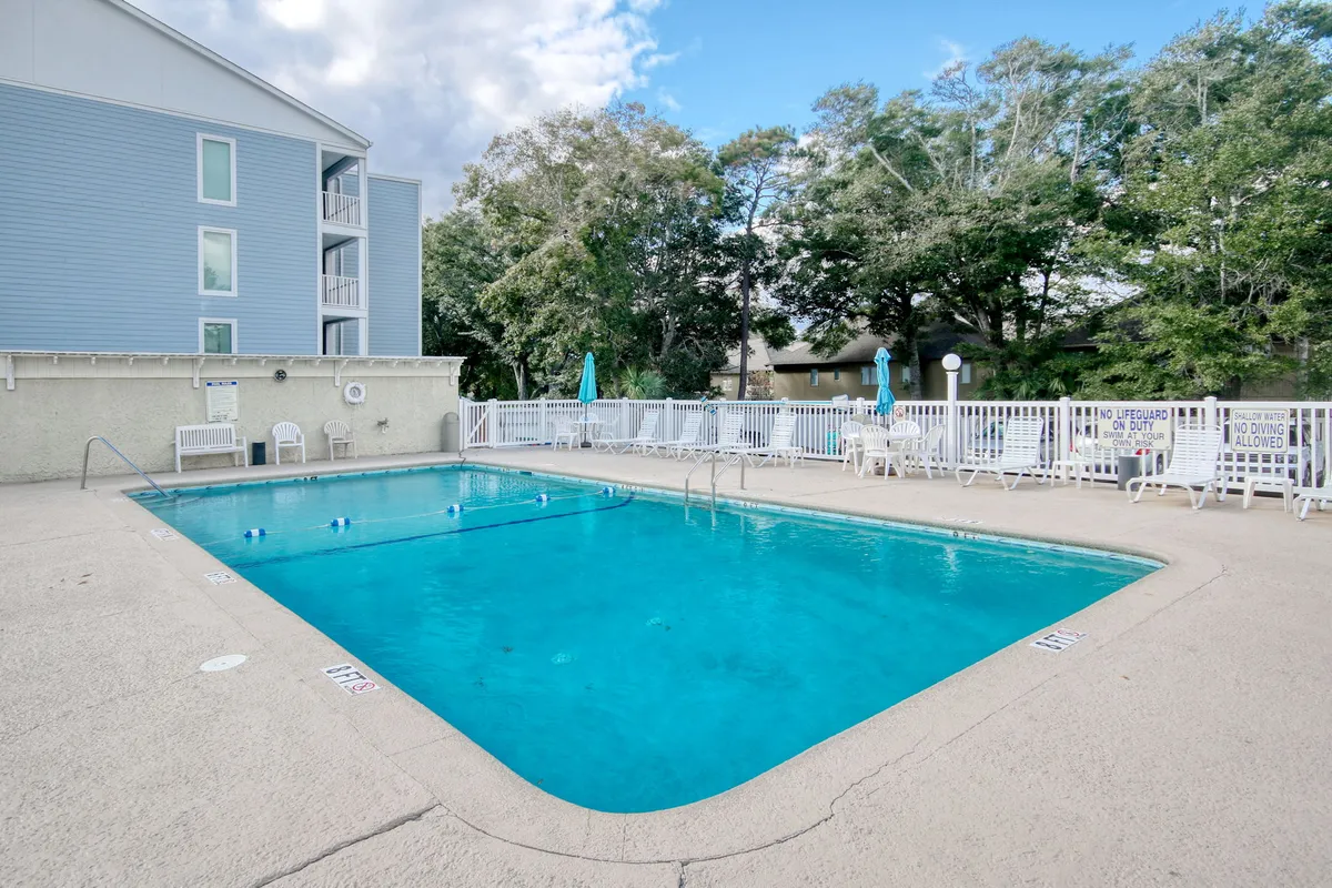 This exterior image features the property's swimming pool surrounded by lush greenery and lounge chairs, enclosed by a poolside fence. The inviting scene is designed for swimming, sunbathing, and relaxation, offering a refreshing retreat for families and groups after a day at the beach.