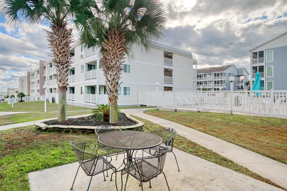 An inviting outdoor seating area framed by palm trees, with a table and chairs set near a white building. In the background, the pool area is visible, reinforcing the connection between communal spaces and outdoor living. The scene communicates relaxation, group gatherings, and sunny coastal ambiance.