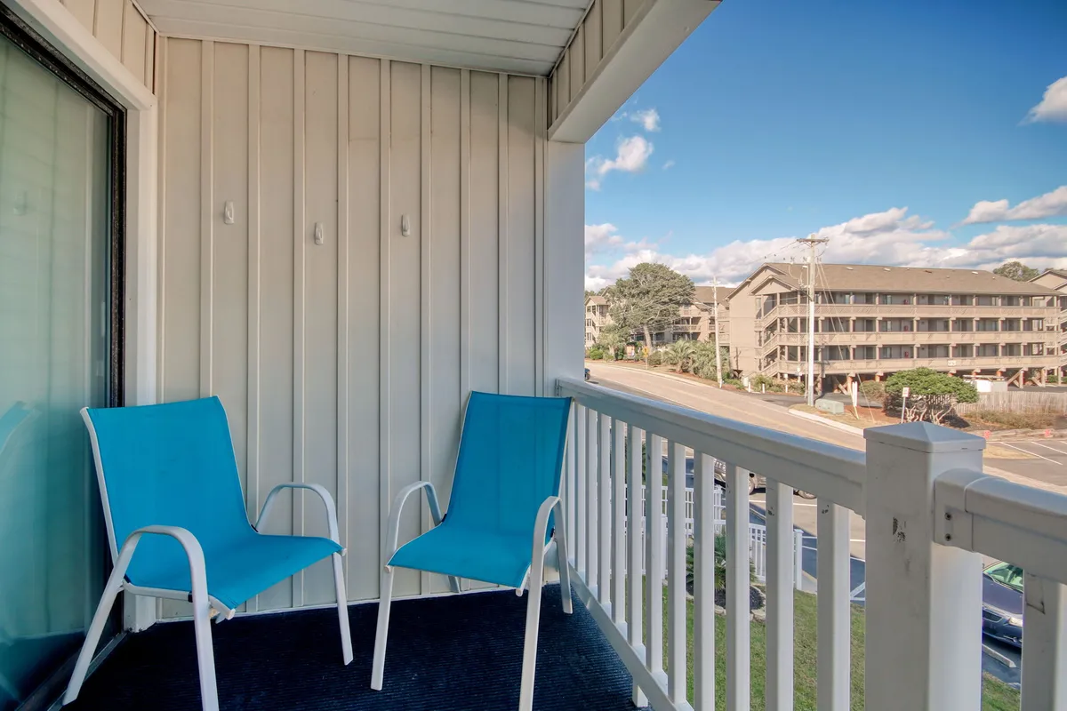 A tranquil balcony scene with two blue chairs positioned for relaxation, a sturdy railing, and a view overlooking the street below. The setup invites moments of quiet reading, sipping drinks, or watching the sunset in comfort and peace.