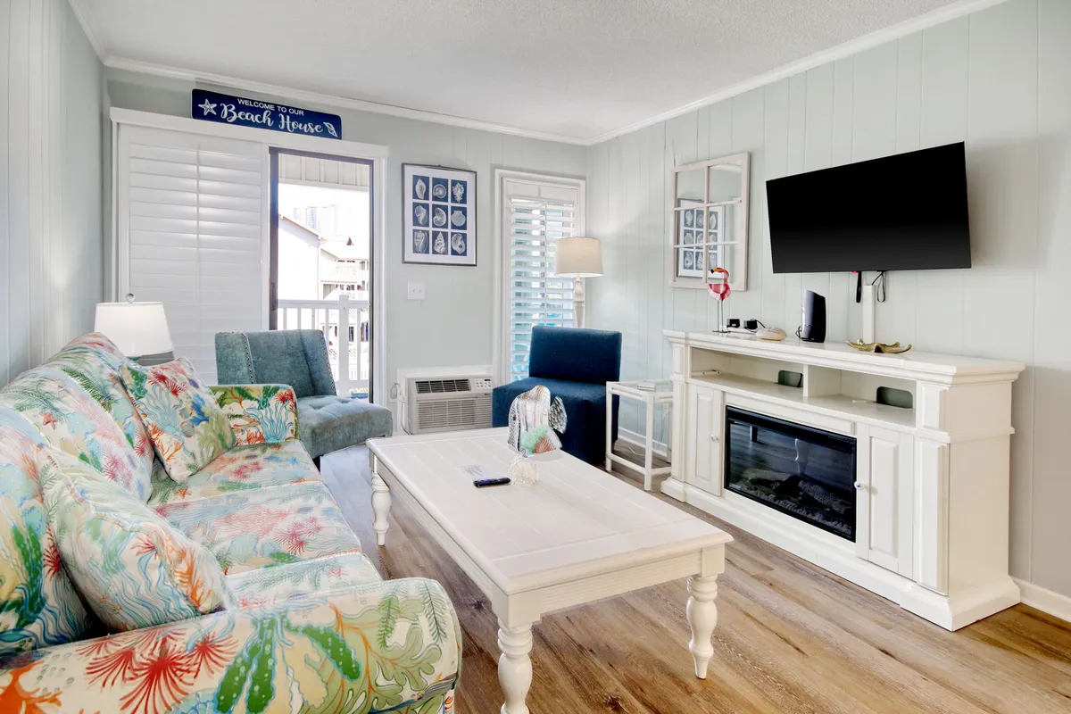 A cheerful living room featuring a colorful sofa, a coffee table, and a TV unit in front of a large window with a view. The space feels spacious and bright, capturing the essence of a coastal gathering spot ideal for relaxing, socializing, or enjoying movie nights.
