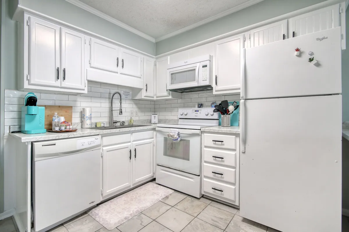 This image showcases a welcoming kitchen space with white cabinetry, stainless steel appliances, a tile backsplash, a kitchen sink, and a small rug. The scene includes hints of cooking and dining activity, suggesting a warm, lively atmosphere perfect for shared meals and socializing.