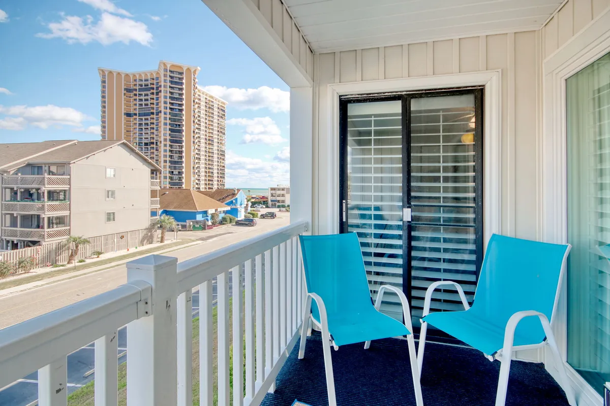 A second-floor balcony with blue chairs facing a sturdy railing, overlooking distant buildings. The space is casual and inviting, ideal for relaxing or soaking in the surroundings, and the elevated perspective hints at ocean breezes and peaceful seclusion.