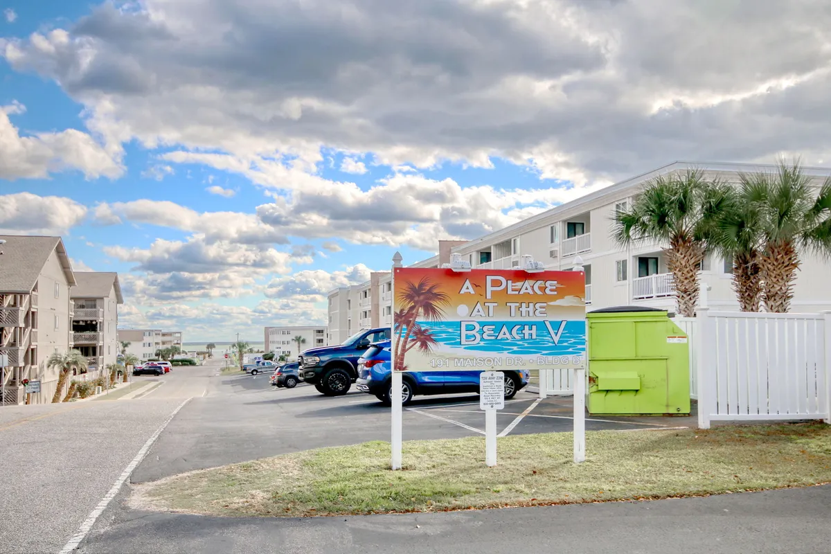 The image features the exterior of the condo building framed by swaying palm trees, with blue skies and soft clouds overhead. Prominent signage welcomes guests, and the relaxed ambiance is enhanced by vehicles and the inviting setting, hinting at easy beach access and vibrant surroundings.